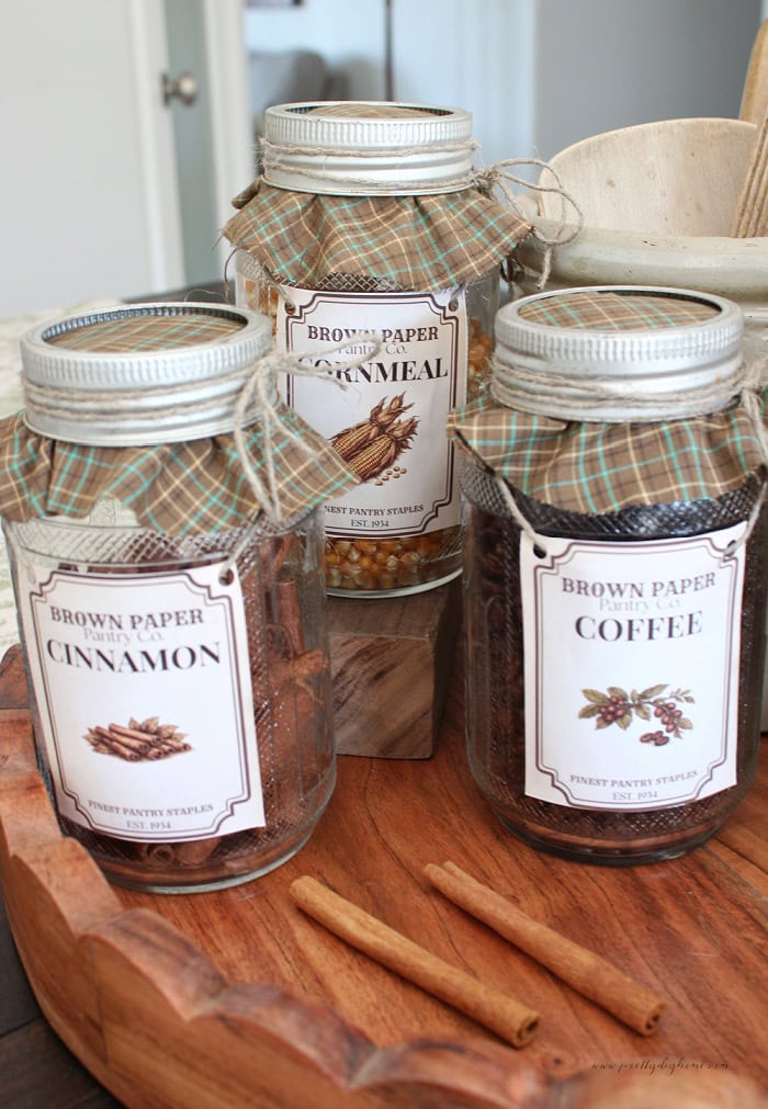 Three vintage glass pantry jars with cinnamon, cornmeal, and coffee labels are finished with plaid fabric lids and twine on a wooden tray.