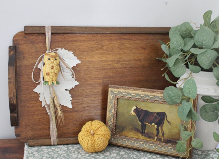 Vintage wood tray decorated for fall with an owl, cream leaves, wheat, pumpkin, framed cow print, and greenery.