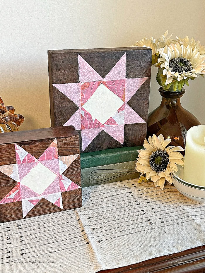 Two handmade wooden barn quilt shelf sitters displayed on vintage books with cream flowers and a candle.