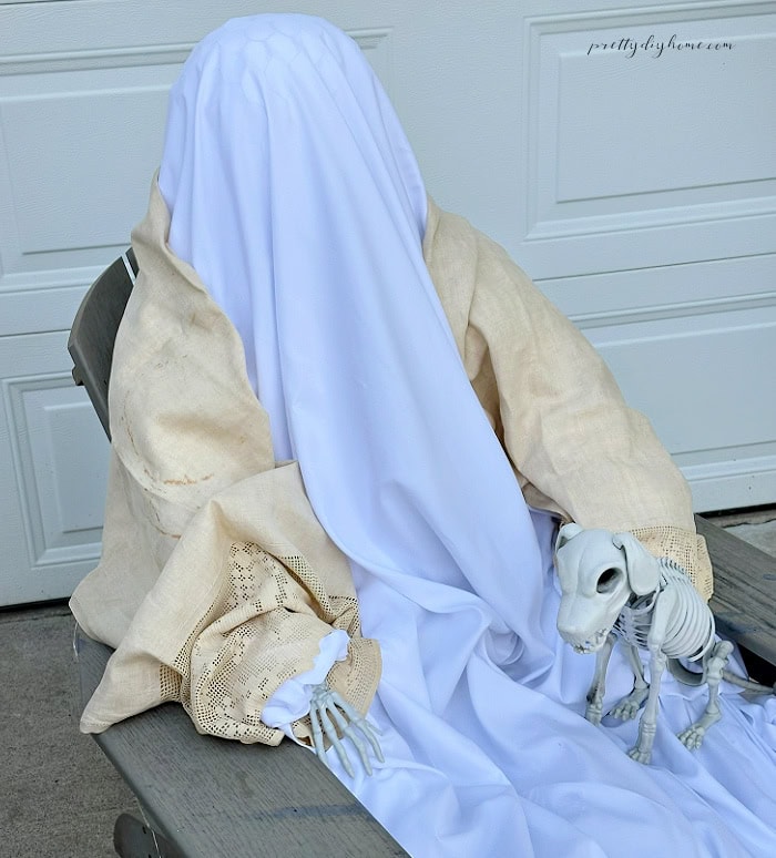 A DIY Halloween ghost is covered with white fabric and a cream vintage shawl beside a small skeleton dog.