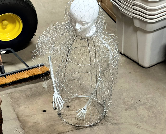 The upper half of a DIY Halloween ghost body is formed with chicken wire, wire arms, and a styrofoam head.