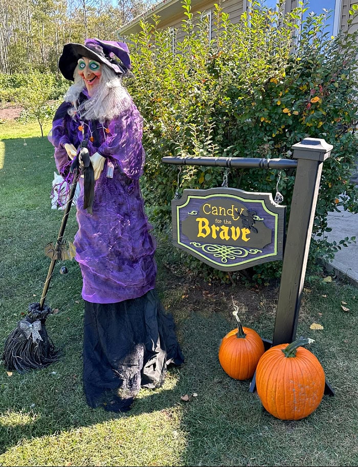 DIY Halloween sign displayed in the front yard with a witch decoration and orange pumpkins.