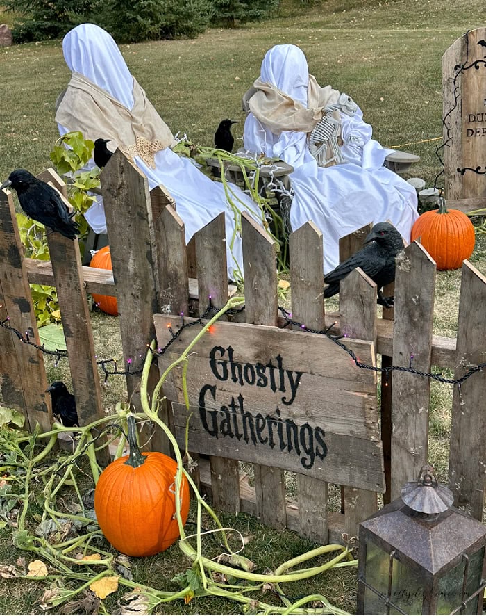 Two handmade outdoor Halloween ghosts are displayed behind a Ghostly Gatherings sign with pumpkins, crows, and rustic pallet decorations.
