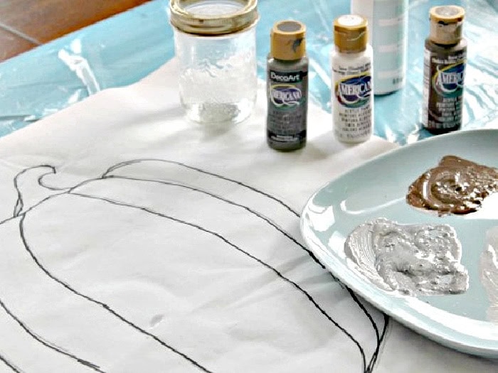 Supplies for painting a DIY pumpkin pillow including acrylic paint, fabric medium, brushes, and a pumpkin template.