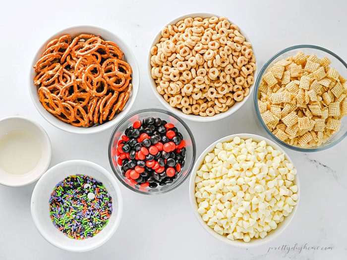 Ingredients for an easy Halloween party snack mix including Rice Chex, Honey Nut Cheerios, pretzels, white chocolate chips, Halloween M&Ms, and sprinkles.