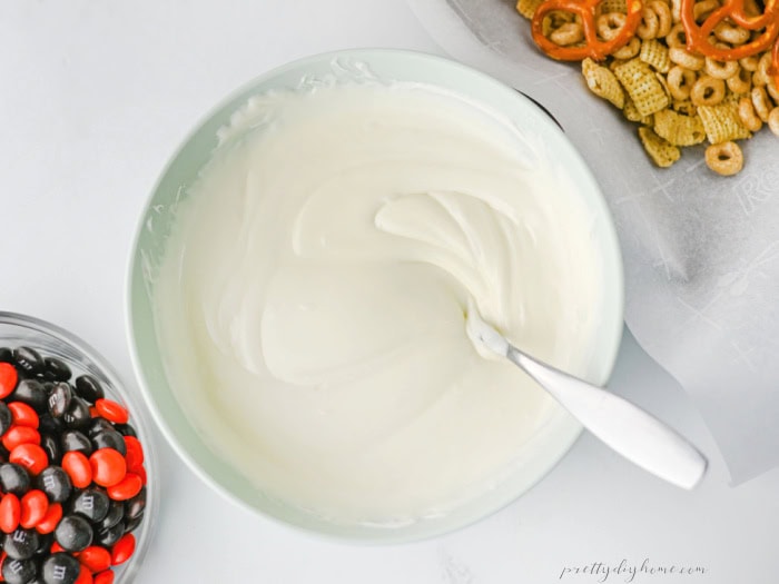 Melted white chocolate for an easy Halloween party snack mix in a bowl beside the cereal and candy ingredients.