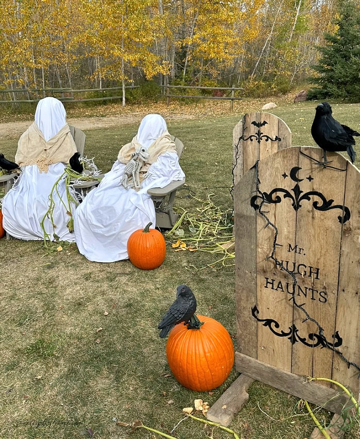 Two handmade outdoor Halloween ghosts sit beside the Mr. Hugh Haunts pallet gravestone in a rustic Halloween yard display with pumpkins and crows.