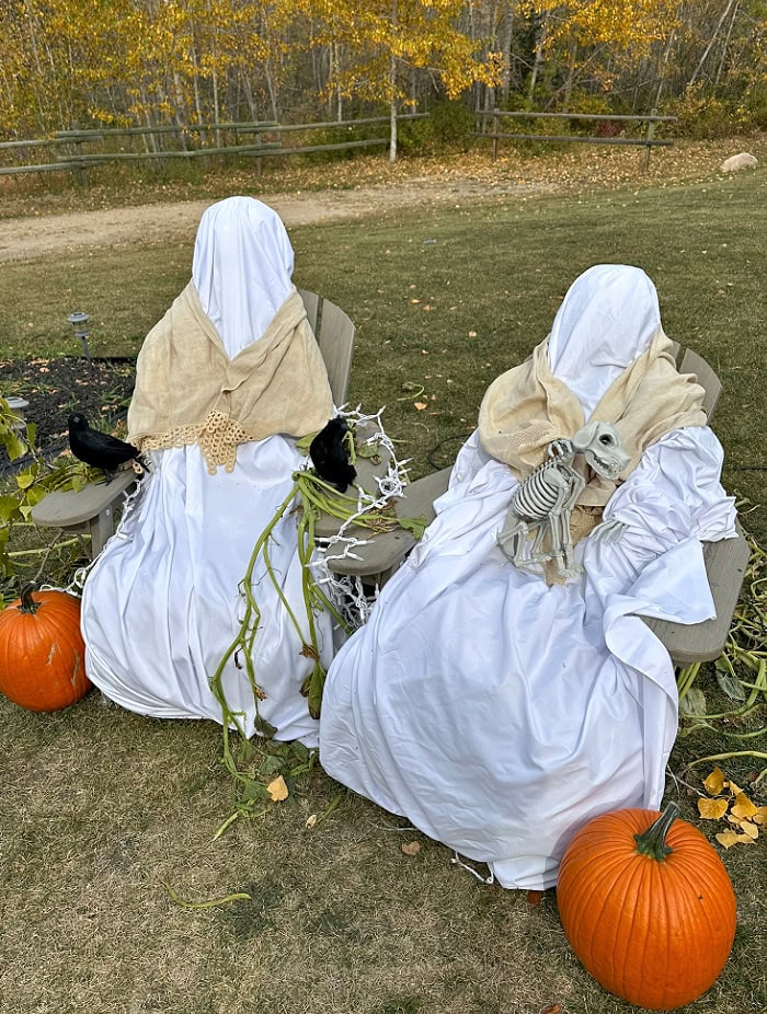Two primitive Halloween ghosts sit outdoors in Adirondack chairs with white fabric, cream shawls, pumpkins, crows, and a small skeleton dog.