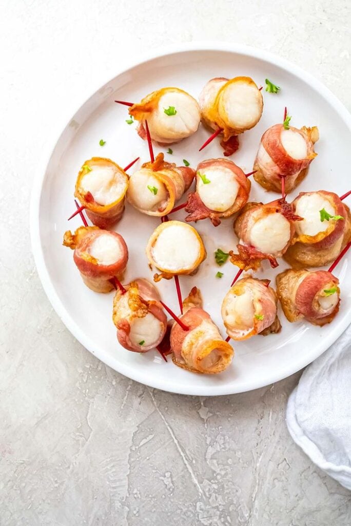 Air fryer bacon-wrapped scallops cooked until golden, secured with toothpicks, and garnished with fresh parsley on a white serving plate.