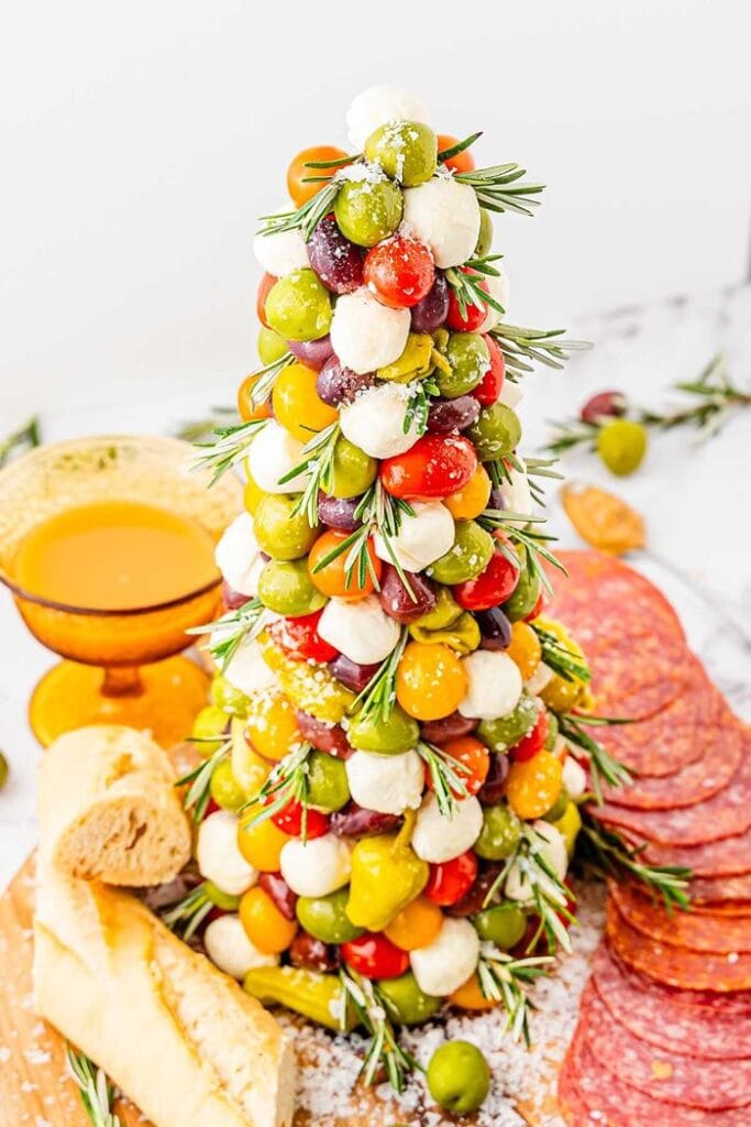 Antipasto Christmas tree decorated with salami, mozzarella cheese balls, cherry tomatoes, green olives, and fresh herbs arranged in a festive tree shape.