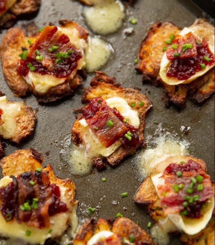 Crispy smashed potatoes topped with melted brie cheese, cranberry sauce, bacon, and fresh chives on a baking tray.