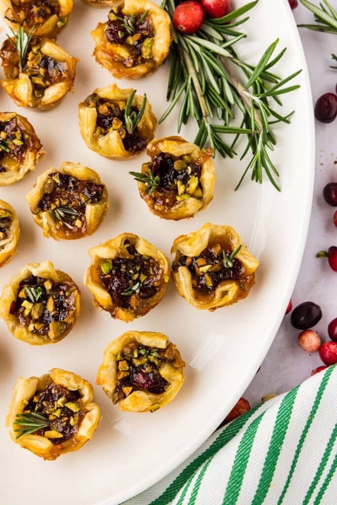 Golden puff pastry brie bites filled with apricot jam, dried cranberries, and chopped pistachios, garnished with fresh rosemary.