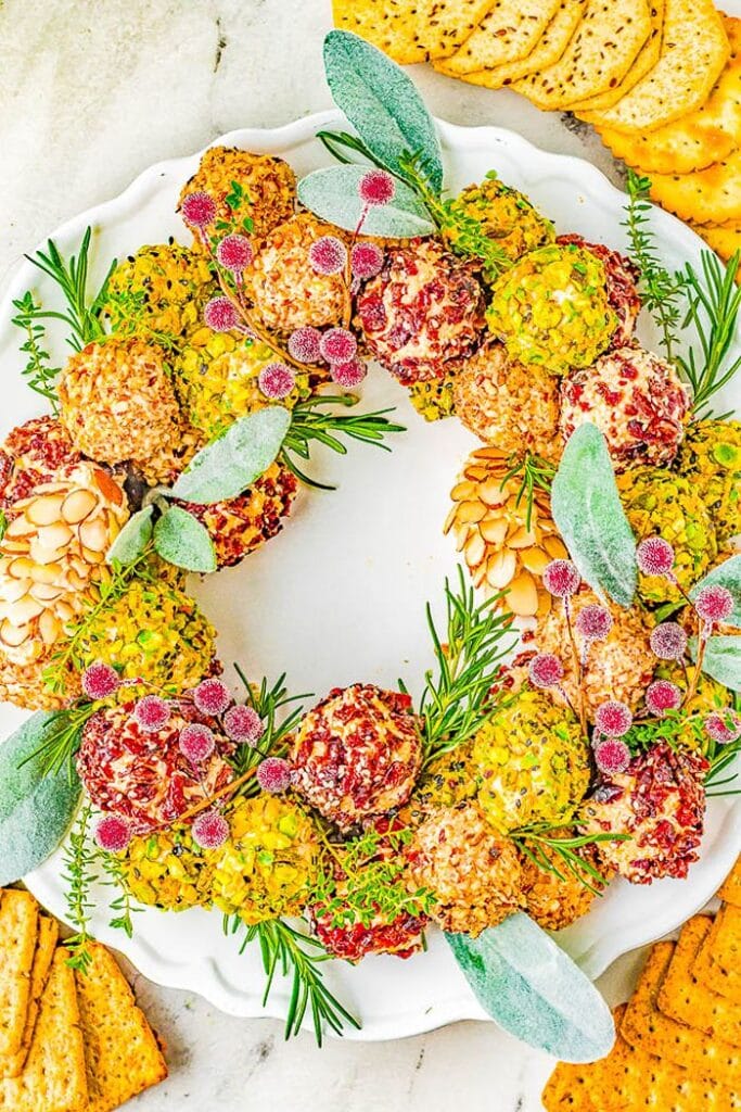 Christmas cheese ball wreath made with bite-sized cheese balls coated in nuts, herbs, and cranberries, arranged with fresh greenery on a white platter.