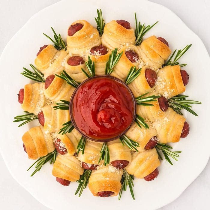 Christmas pigs in a blanket wreath made with golden pastry-wrapped sausages, fresh rosemary sprigs, and a bowl of dipping sauce in the centre.