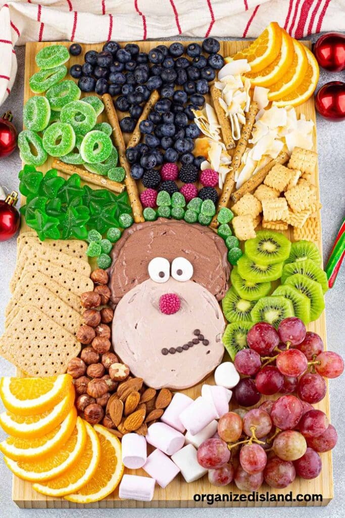 Christmas reindeer charcuterie board featuring a reindeer face surrounded by fresh fruit, crackers, nuts, cheese, marshmallows, and colourful Christmas candies.