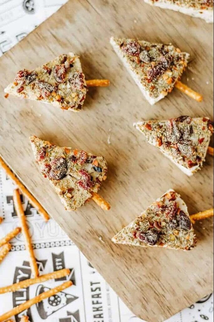 Mini Christmas tree cheese appetizers made with triangular cheese wedges coated in herbs, nuts, and cranberries, with pretzel sticks for trunks.