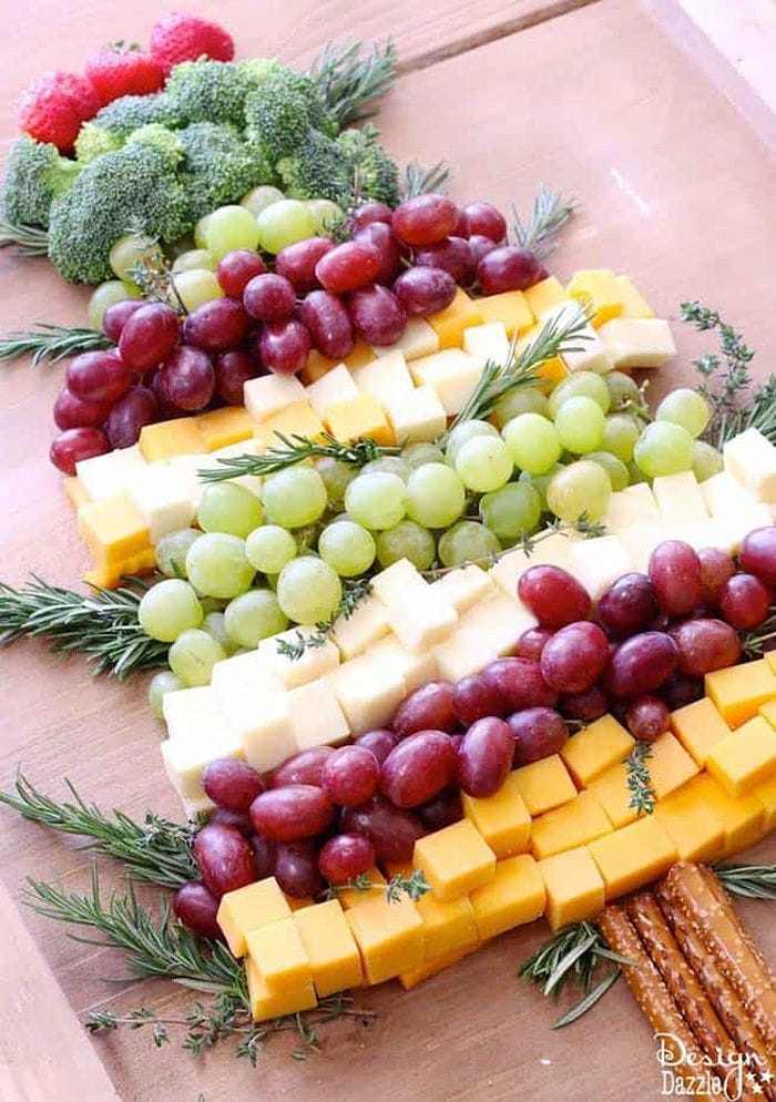 Christmas tree appetizer tray arranged with cheddar and white cheese cubes, red and green grapes, broccoli, strawberries, and fresh rosemary on a wooden board.