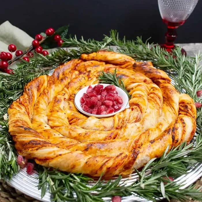 Golden twisted pizza wreath made with flaky pastry and pizza filling, arranged in a circle with marinara dipping sauce in the centre.