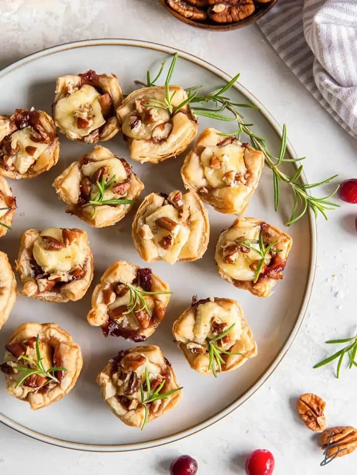 Golden puff pastry cranberry brie bites topped with melted brie cheese, cranberry sauce, chopped pecans, and fresh rosemary.
