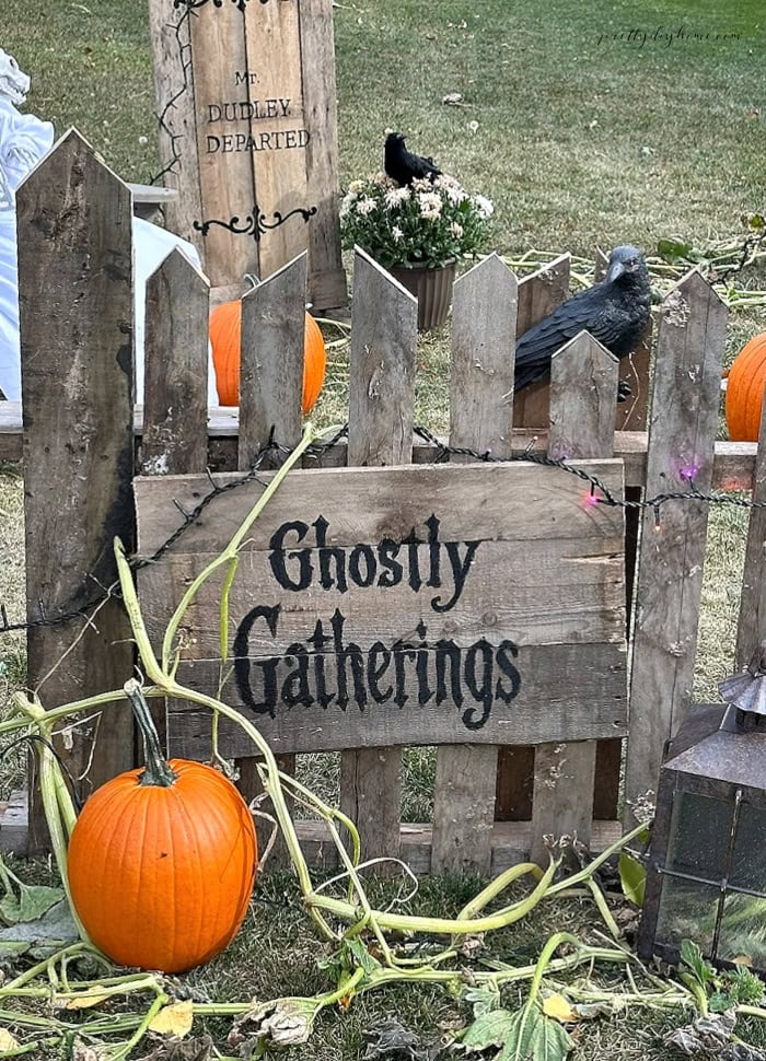 DIY Halloween pallet fence with a Ghostly Gatherings sign, pumpkin, raven, gravestone, vines, and string lights.