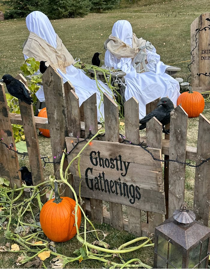 Ghostly Gatherings Halloween yard display with a DIY pallet fence, seated ghosts, ravens, pumpkins, vines, and lantern.