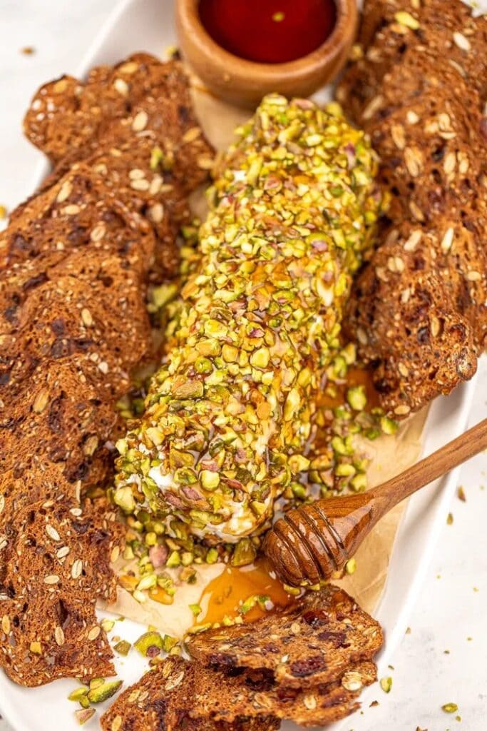 Goat cheese log coated in chopped pistachios and drizzled with hot honey, served with crisp crackers on an appetizer platter.