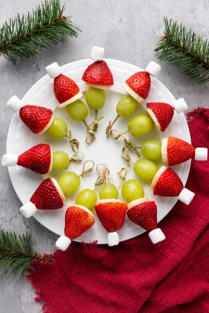 Grinch fruit kabobs made with green grapes, banana slices, strawberries, and mini marshmallows arranged to look like little Santa hats.