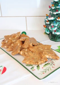 Homemade mixed nut peanut brittle piled on a vintage holly platter beside a ceramic Christmas tree and green ornaments.