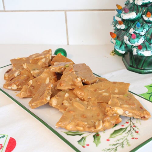 Homemade mixed nut peanut brittle piled on a vintage holly platter beside a ceramic Christmas tree and green ornaments.