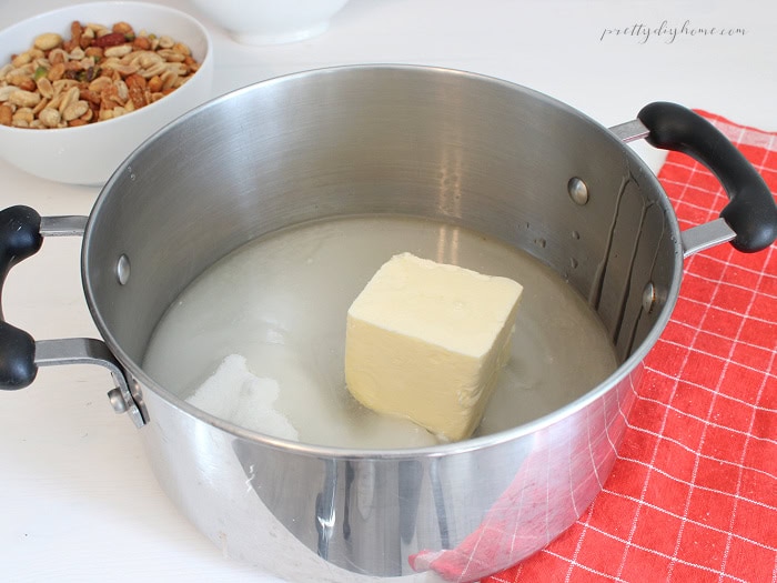 Butter and sugar in a stainless steel saucepan with mixed nuts in the background, ready to make homemade peanut brittle.