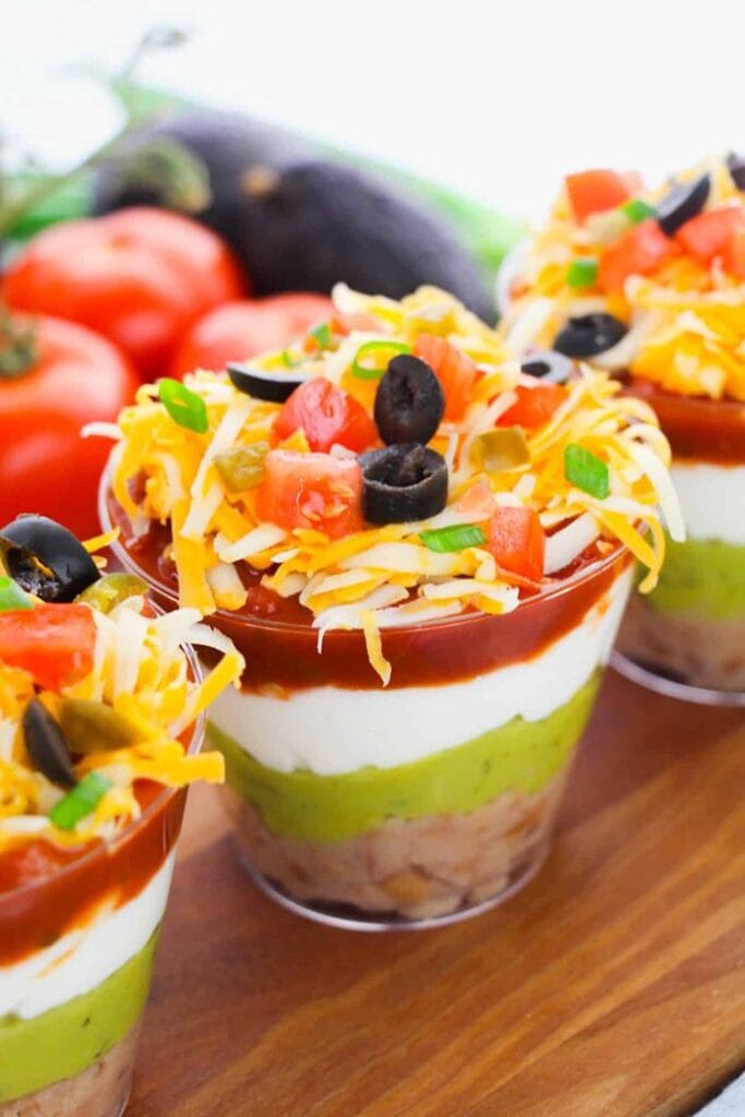 Individual seven-layer dip cups filled with refried beans, guacamole, sour cream, salsa, shredded cheese, black olives, tomatoes, and green onions.