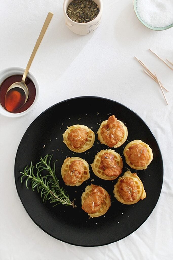 Mini chicken and waffles topped with crispy chicken bites, fresh herbs, and maple syrup, arranged on a black serving platter.