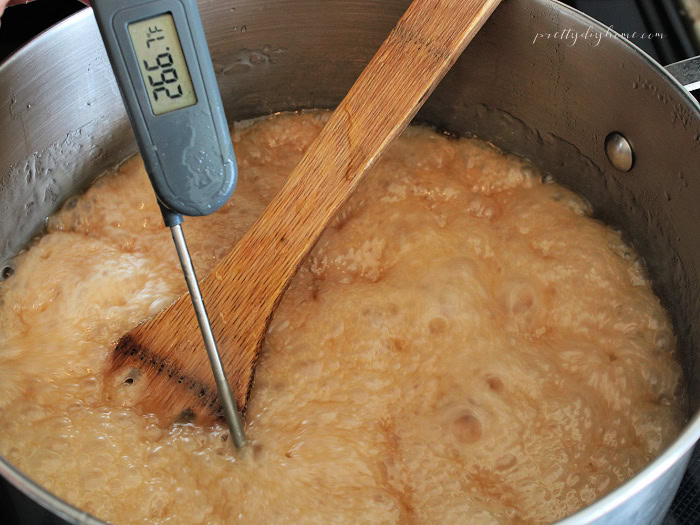 Golden sugar mixture boiling at 267°F in a stainless steel saucepan with a candy thermometer and wooden spoon while making homemade mixed nut brittle.
