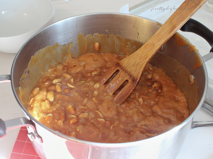 Mixed nuts being stirred into golden homemade peanut brittle in a stainless steel saucepan with a wooden spoon.