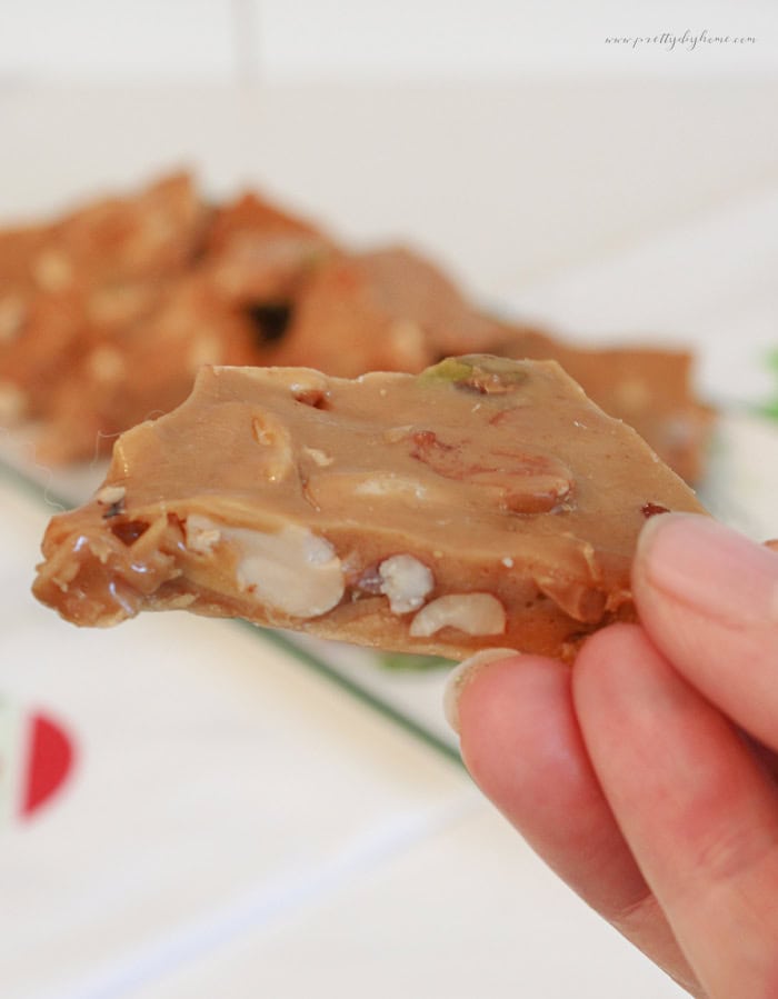 Close-up of a thick piece of homemade mixed nut peanut brittle held between fingers, showing crunchy golden candy packed with assorted nuts.