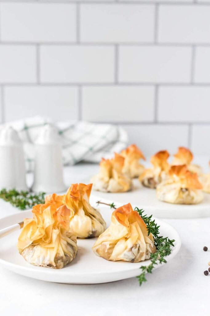Golden, crispy filo pastry parcels filled with mushrooms and goat cheese, arranged on white serving plates with fresh thyme.