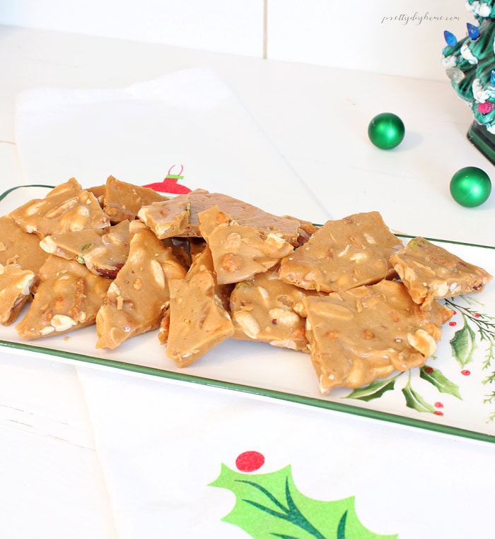Old-fashioned mixed nut peanut brittle piled on a vintage holly platter, showing thick golden pieces filled with peanuts, cashews, almonds, and pistachios.