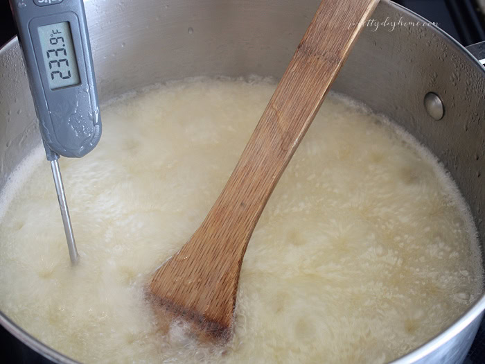 Sugar mixture boiling at 234°F in a stainless steel saucepan with a candy thermometer and wooden spoon while making homemade peanut brittle.