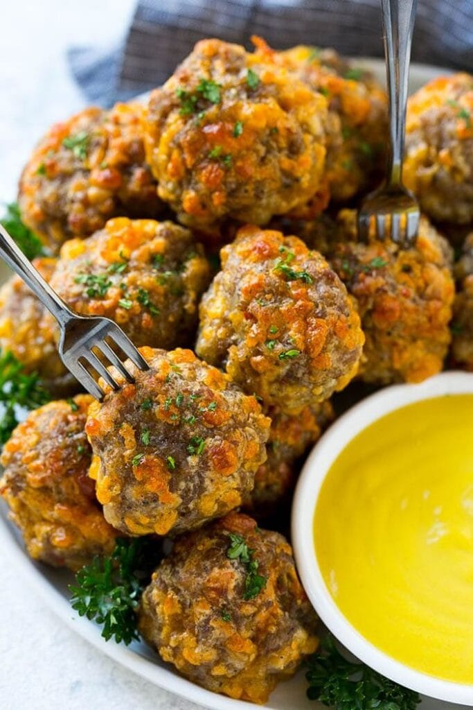 Golden baked sausage cheese balls sprinkled with fresh parsley and served with mustard dipping sauce on a white platter.