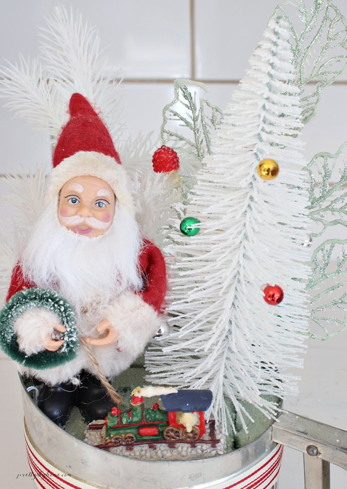 Vintage Christmas flour sifter arrangement featuring a miniature Santa Claus, white bottle brush tree, sparkly green picks, and a tiny Christmas train.