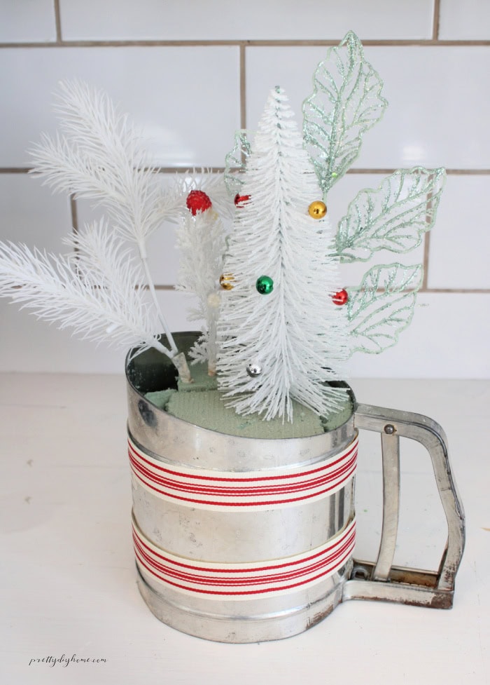 Vintage flour sifter Christmas arrangement featuring a white bottle brush tree, vintage white Christmas picks, and sparkly green greenery arranged in floral foam.