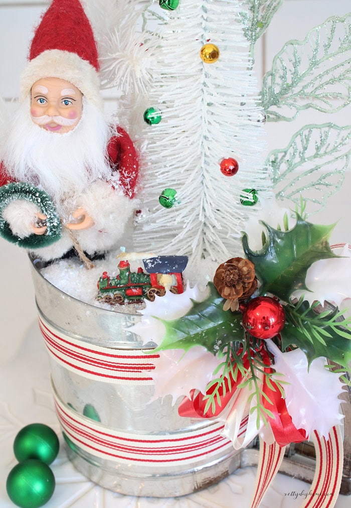 Close-up of vintage Christmas kitchen decorations featuring a Santa Claus, white bottle brush tree, miniature Christmas train, green ornaments, and faux snow in an old flour sifter.