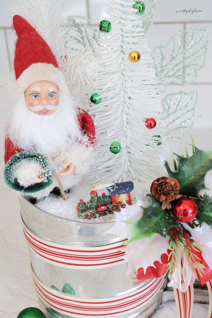 Finished vintage Christmas kitchen arrangement featuring Santa Claus, a white bottle brush tree, miniature train, green bells, red poinsettia, and faux snow in an old metal flour sifter.