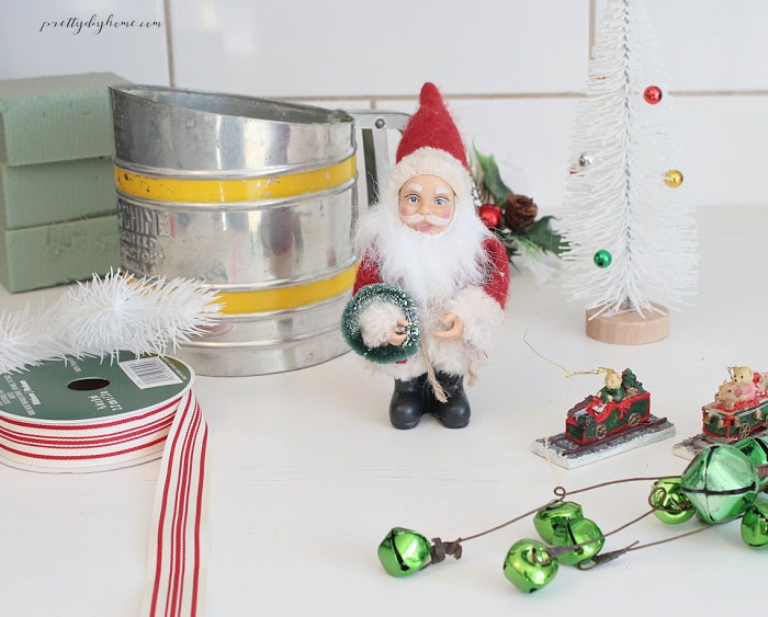 Supplies for DIY vintage Christmas kitchen decorations, including an old flour sifter, miniature Santa Claus, white bottle brush tree, ticking ribbon, vintage ornaments, and Christmas picks.