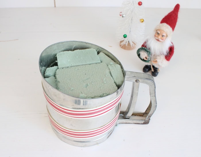 Vintage metal flour sifter filled with floral foam and decorated with red and white ticking ribbon, preparing the container for a DIY Christmas kitchen arrangement.