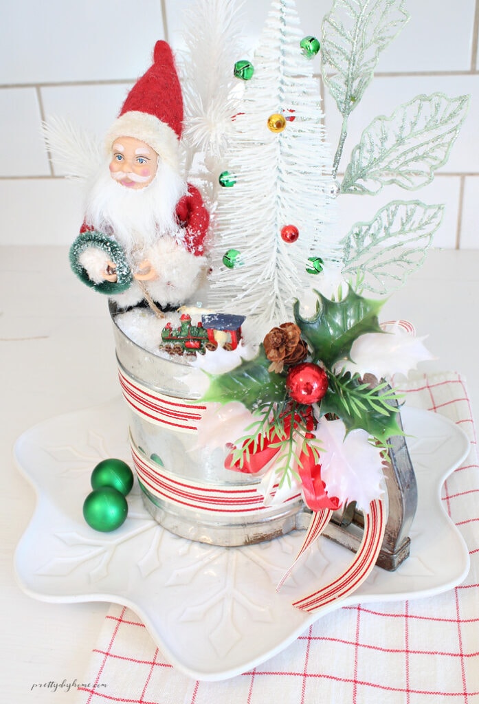 Vintage Christmas kitchen decorations featuring a Santa Claus, white bottle brush tree, miniature train, green ornaments, and faux snow arranged in an antique flour sifter on a white plate.